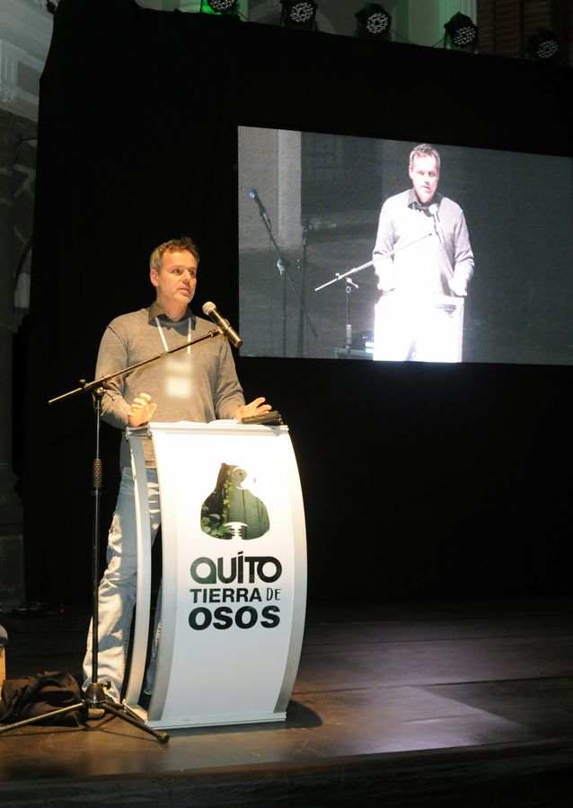 Speaker addresses a conference from a podium, projected live on a large screen