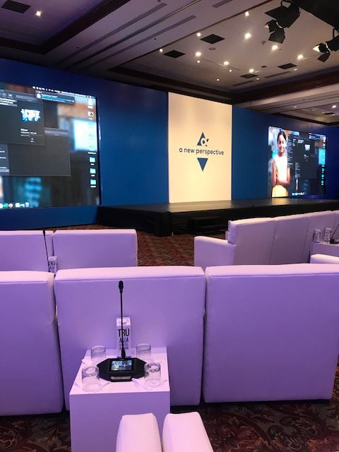 White lounge seating with delegate microphones facing a blue stage and screens