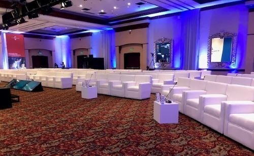 Hotel ballroom arranged with white lounge sofas and blue uplighting for a conference