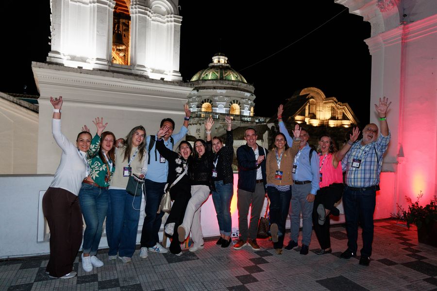Joyful group of delegates waving on a rooftop terrace beside an illuminated colonial bell tower and church domes at night