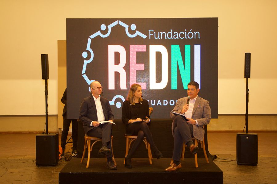 Three panelists seated on a black stage before a large LED screen with foundation branding