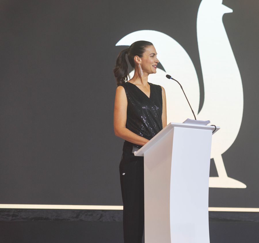 Host in a sequined top smiling at a white podium in front of a large illuminated stage screen