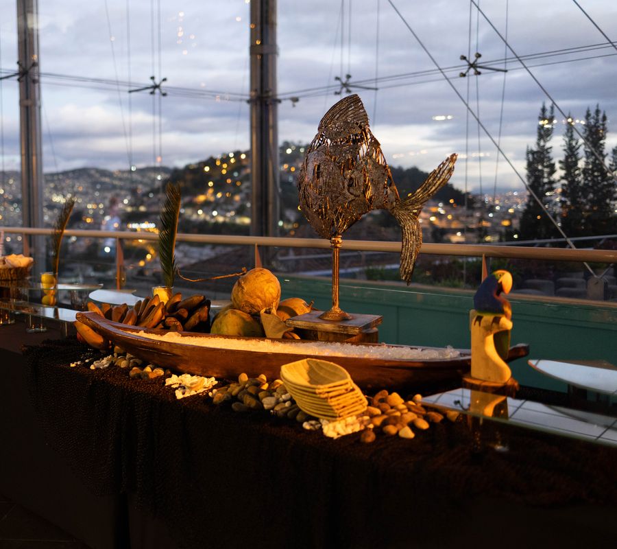 Tropical buffet display with a bronze fish sculpture, a wooden canoe filled with ice, coconuts and plantains overlooking city lights at dusk