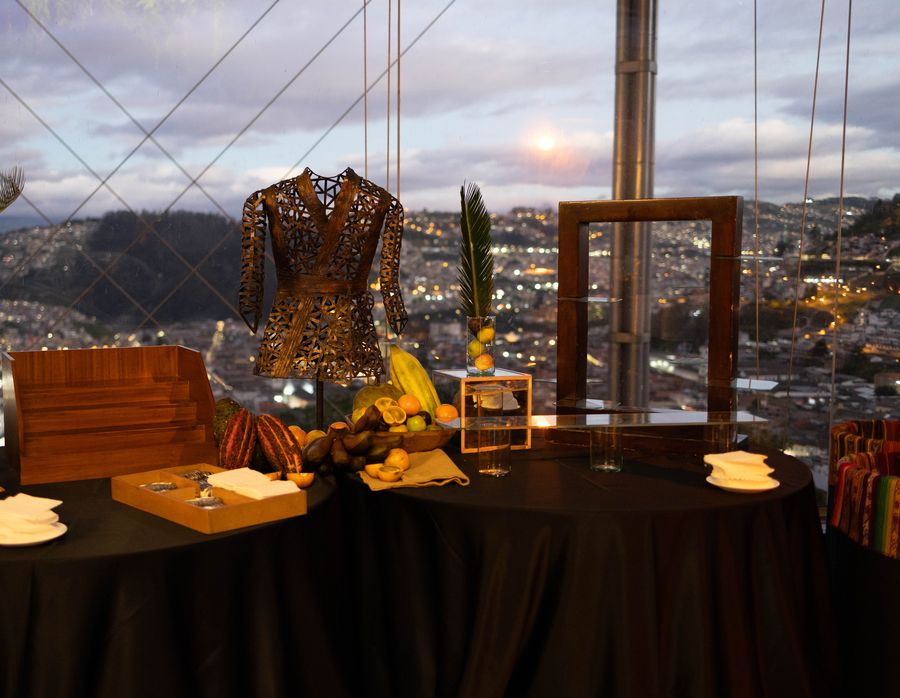 Cacao pods and tropical fruit display beside an openwork metal jacket sculpture, overlooking city lights at dusk