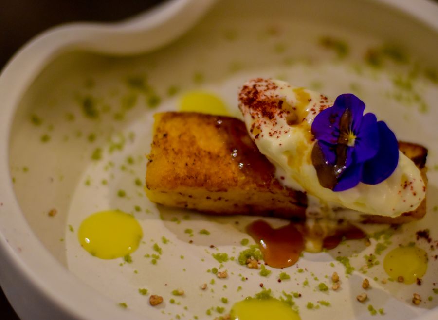 Caramelized brioche-style dessert with a cream quenelle, golden fruit sauce dots, green crumble and an edible purple viola