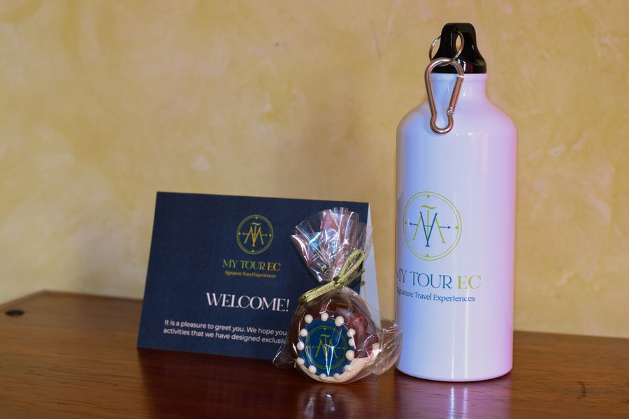 Welcome kit with a white branded water bottle, a navy welcome card and a logo-topped chocolate wrapped in cellophane on a wooden table